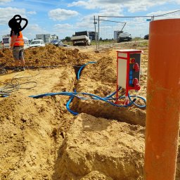 Elgrom - Wykop z ułożonymi niebieskimi rurami kablowymi, skrzynka elektryczna z gniazdami na placu budowy. Pracownik w odblaskowej kamizelce nadzoruje prace. Słoneczny dzień.