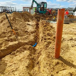 Elgrom - Wykopany rów w piasku z niebieską rurą w środku i pomarańczową rurą obok. W tle koparka i pracownik w kamizelce. Prace ziemne pod instalacje.