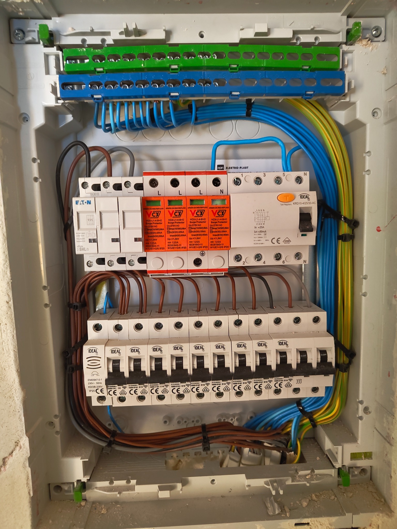 Rozdzielnica elektryczna w obudowie, widoczne zabezpieczenia przepięciowe VCX, wyłączniki IDEAL, przewody w kolorach niebieskim, żółto-zielonym i brązowym, zamocowane w listwach.