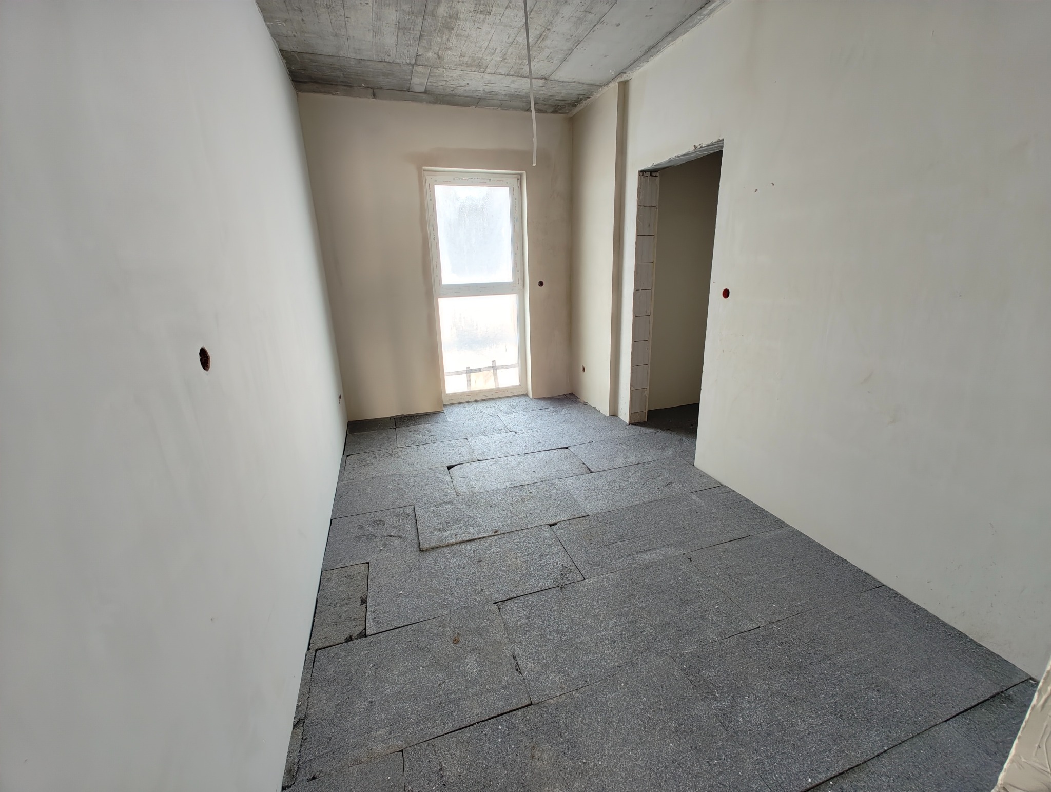 Surowe wnętrze z oknem i szarą izolacją podłogową. Widoczne otwory na gniazdka elektryczne w ścianach. Betonowy sufit bez wykończenia.