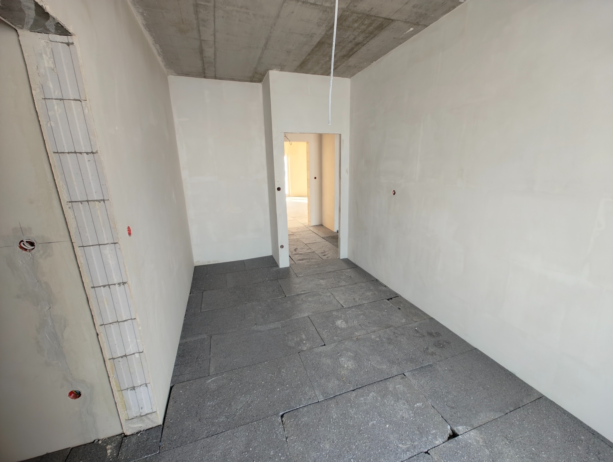 Surowy stan wnętrza: szare płyty podłogowe, gładkie, białe ściany, widoczne punkty elektryczne i wystające przewody. W tle otwarte przejście do kolejnego pomieszczenia.