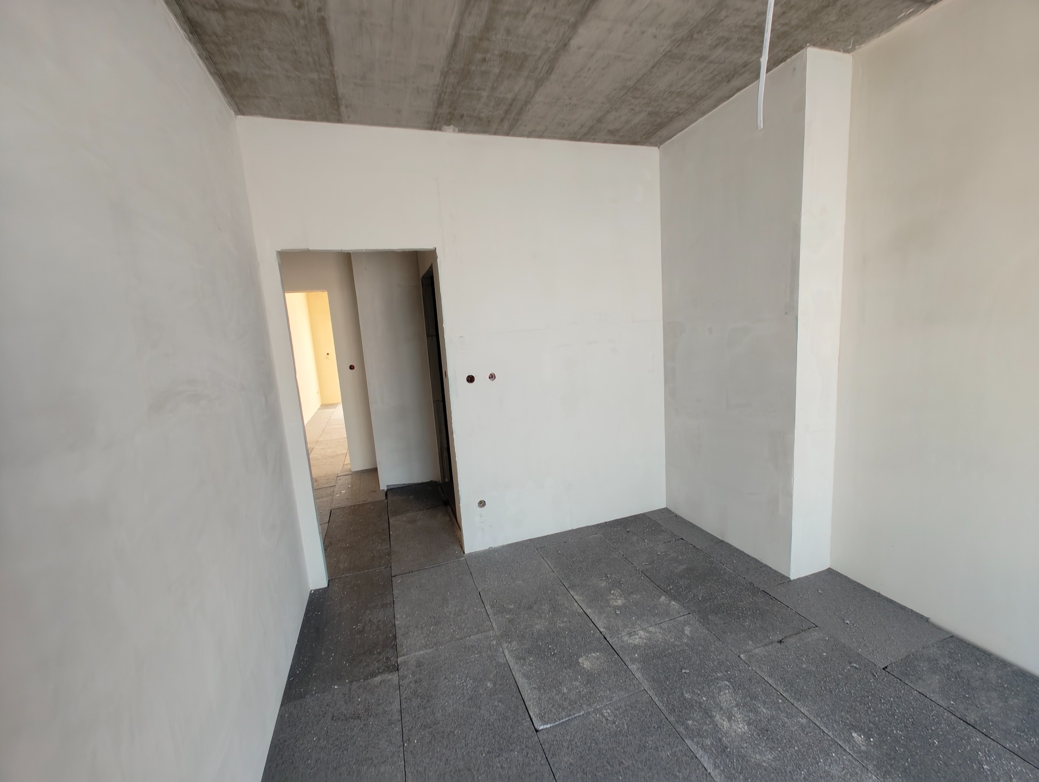 Surowy stan wnętrza: szare płyty podłogowe, białe ściany z widocznymi puszkami elektrycznymi i surowy betonowy strop. Widok na sąsiednie pomieszczenie przez otwarte drzwi.