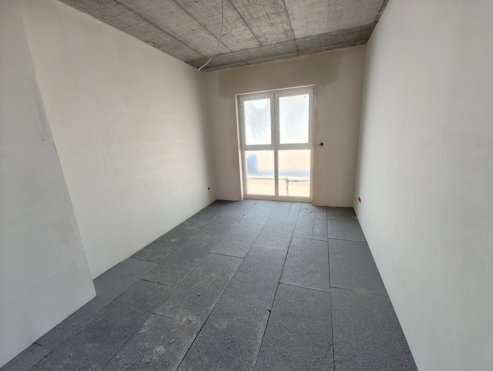 Surowy pokój z betonowym sufitem, przygotowany do prac wykończeniowych. Widoczne kable i puszki elektryczne na ścianach. Podłoga wyłożona płytami izolacyjnymi.