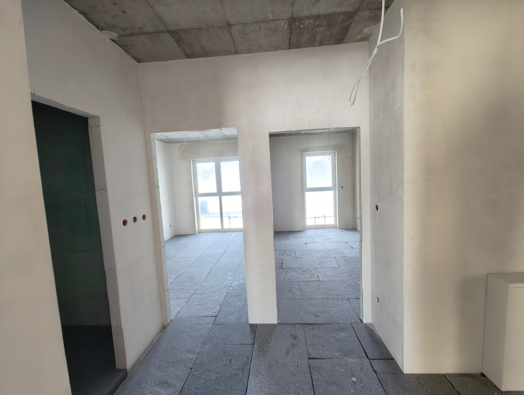 Surowy stan wnętrza z widocznymi przewodami elektrycznymi w ścianach, przygotowany do dalszych prac wykończeniowych. Widoczne okna i surowy betonowy strop.