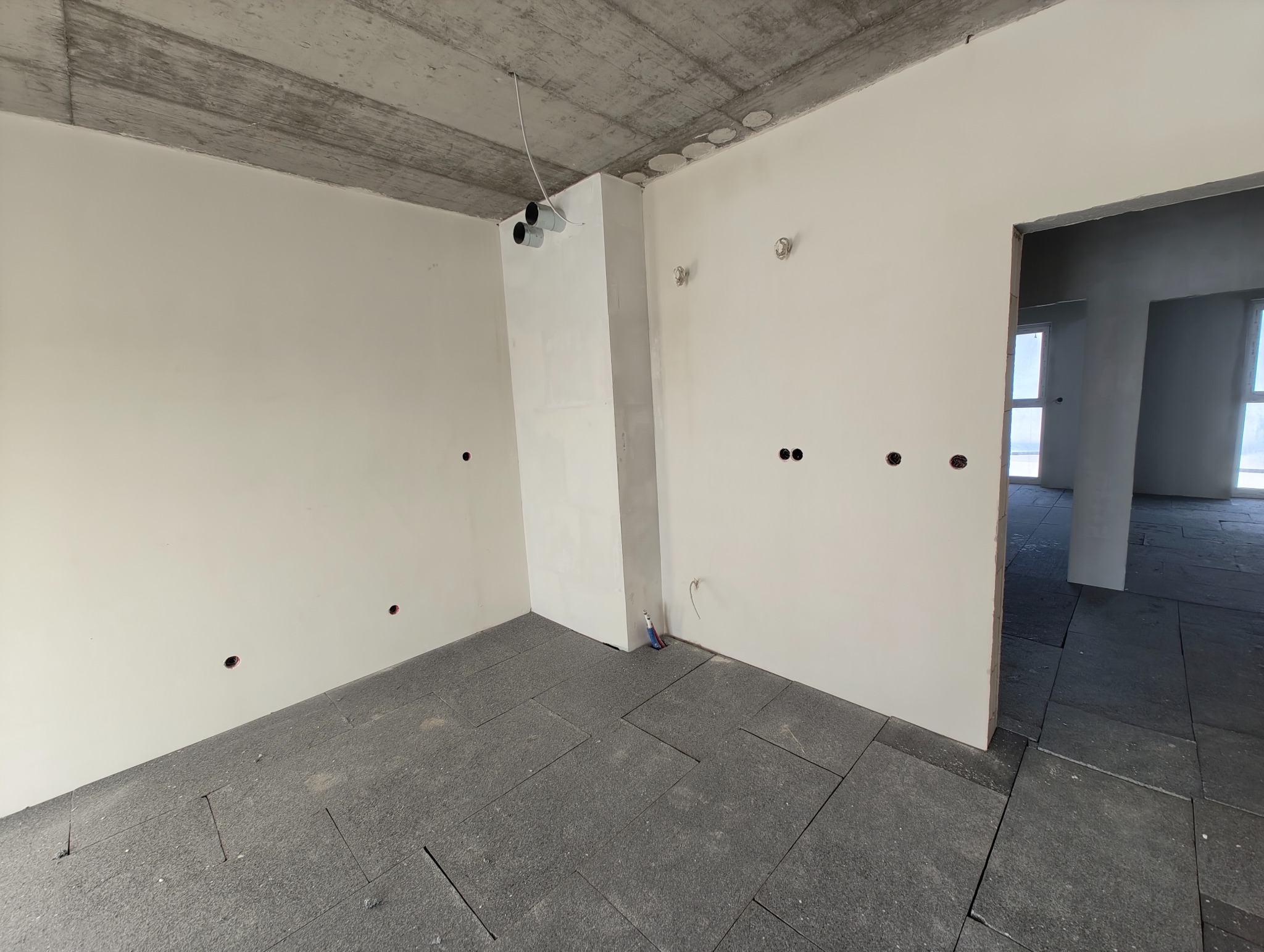 Surowy stan wnętrza: widoczne puszki instalacyjne w ścianach, rury wentylacyjne przy suficie i rozłożona izolacja podłogowa przed dalszymi pracami wykończeniowymi.