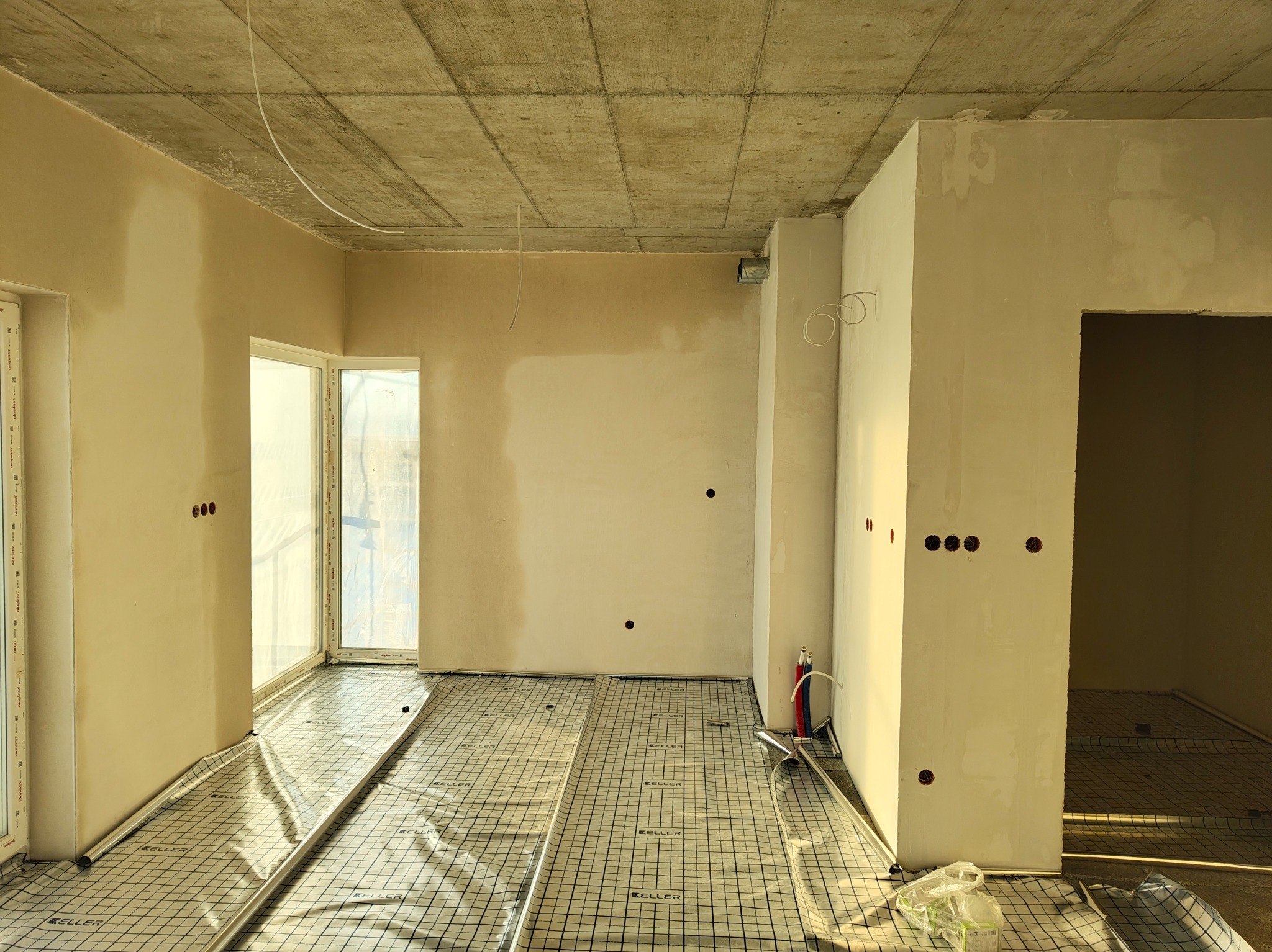 Surowy stan wnętrza z widoczną instalacją ogrzewania podłogowego na folii z nadrukiem, przygotowane otwory pod gniazdka elektryczne w ścianach.