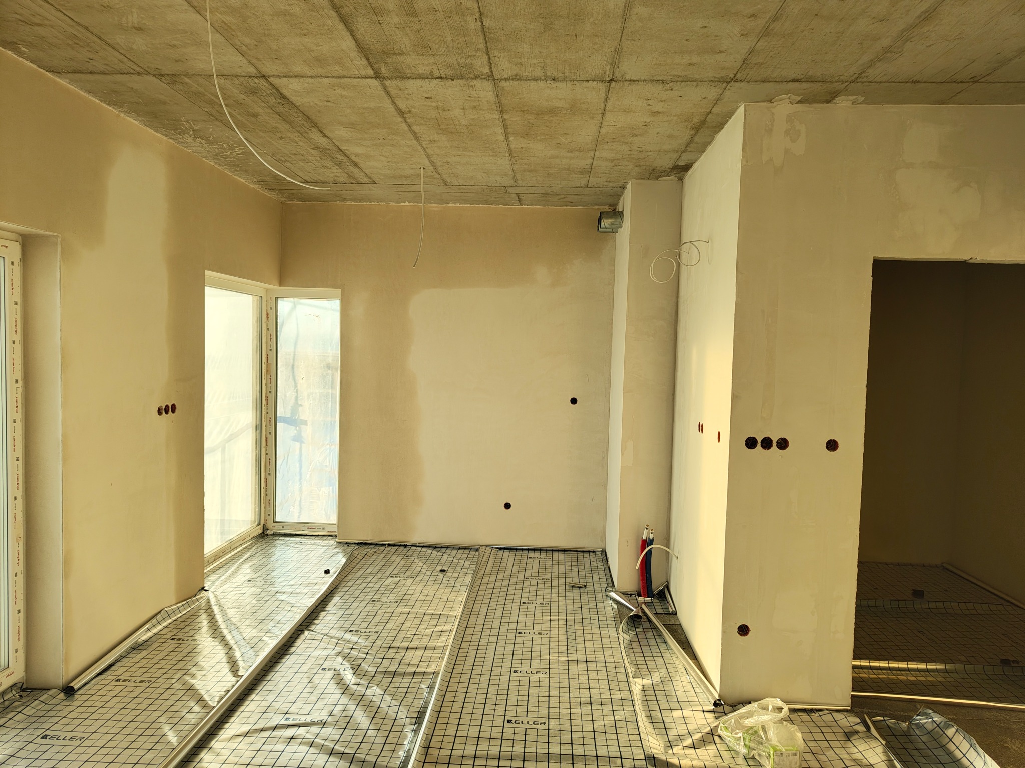 Surowy stan wnętrza z instalacją ogrzewania podłogowego na siatce, widoczne kable elektryczne wystające ze ścian i sufitu, przygotowane pod gniazdka i oświetlenie.