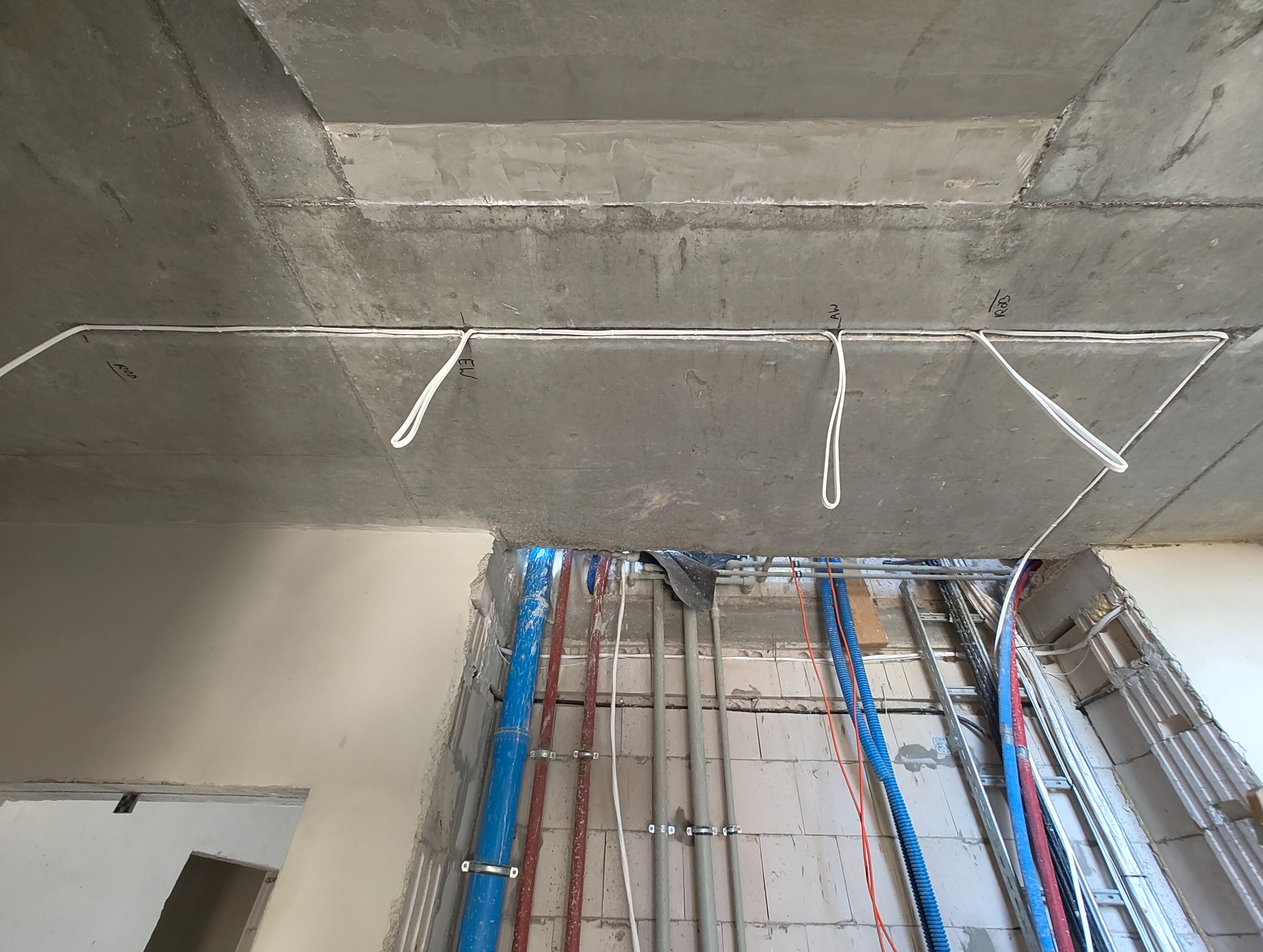 Surowy betonowy strop z poprowadzonymi przewodami elektrycznymi, widoczne oznaczenia. W tle instalacje wodne i kanalizacyjne w trakcie montażu.