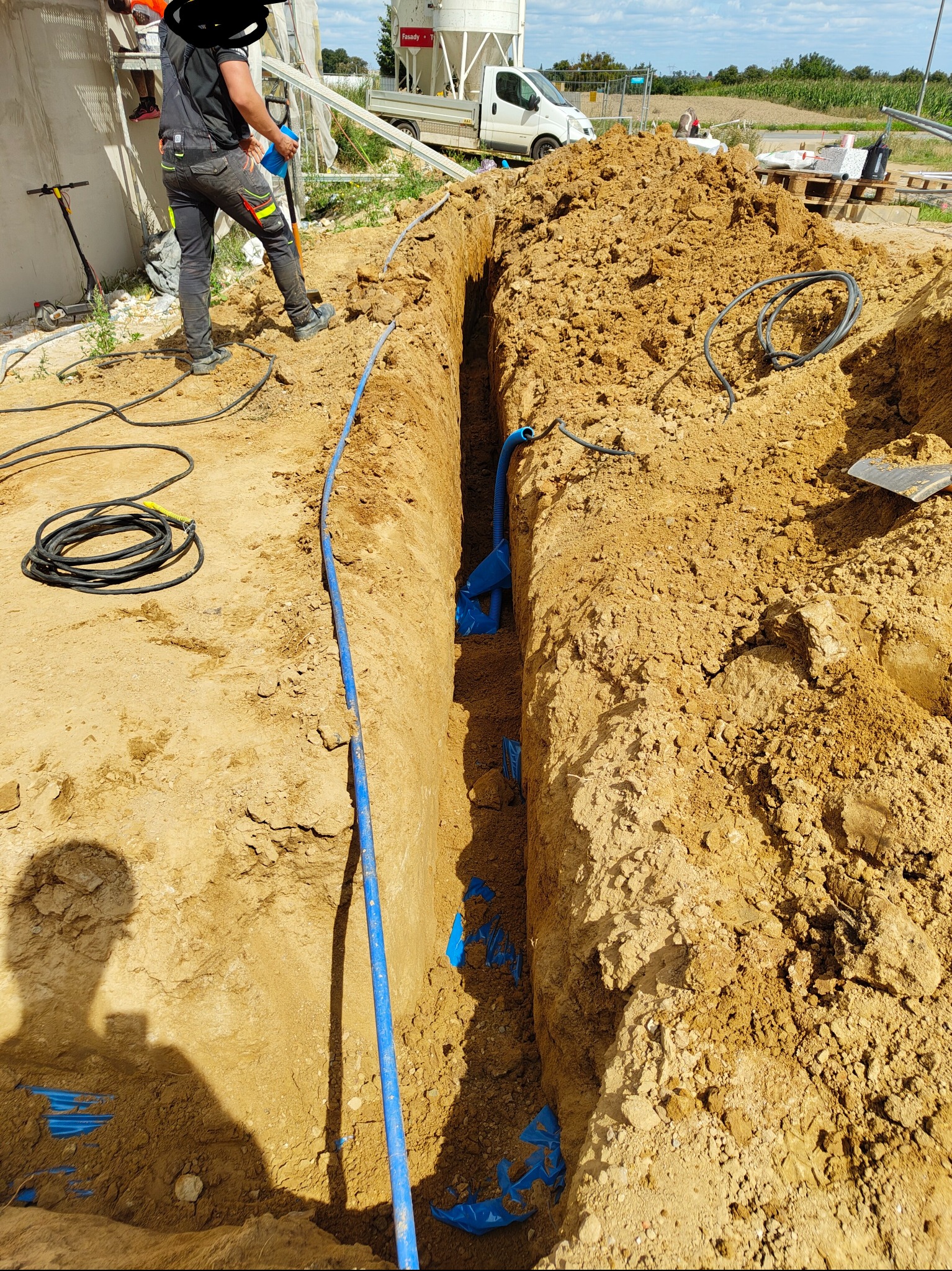 Wykop z ułożonymi niebieskimi rurami kablowymi i czarnym przewodem elektrycznym. Widoczny cień osoby i fragment sylwetki pracownika w kombinezonie.