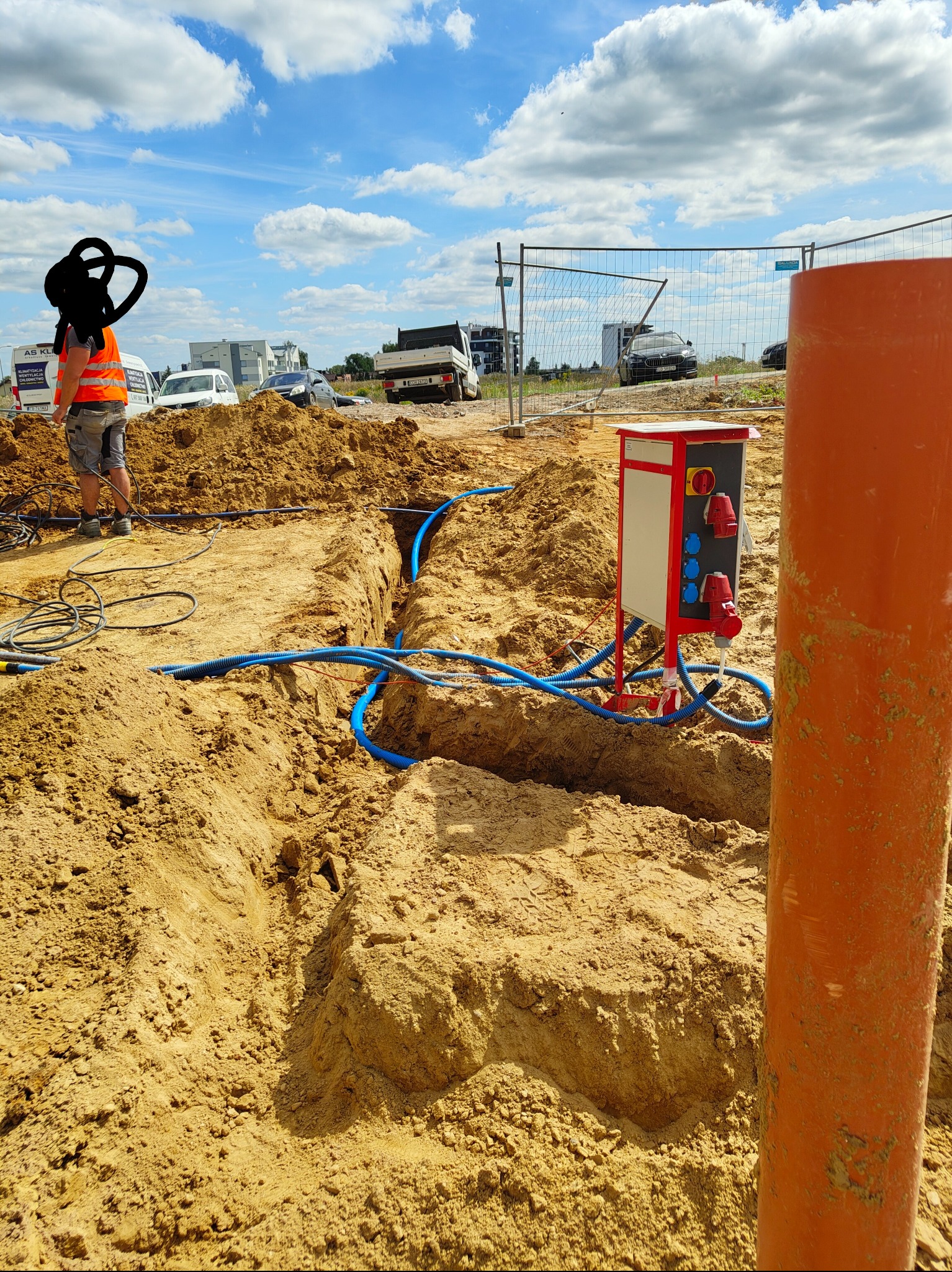 Wykop z ułożonymi niebieskimi rurami kablowymi, skrzynka elektryczna z gniazdami na placu budowy. Pracownik w odblaskowej kamizelce nadzoruje prace. Słoneczny dzień.