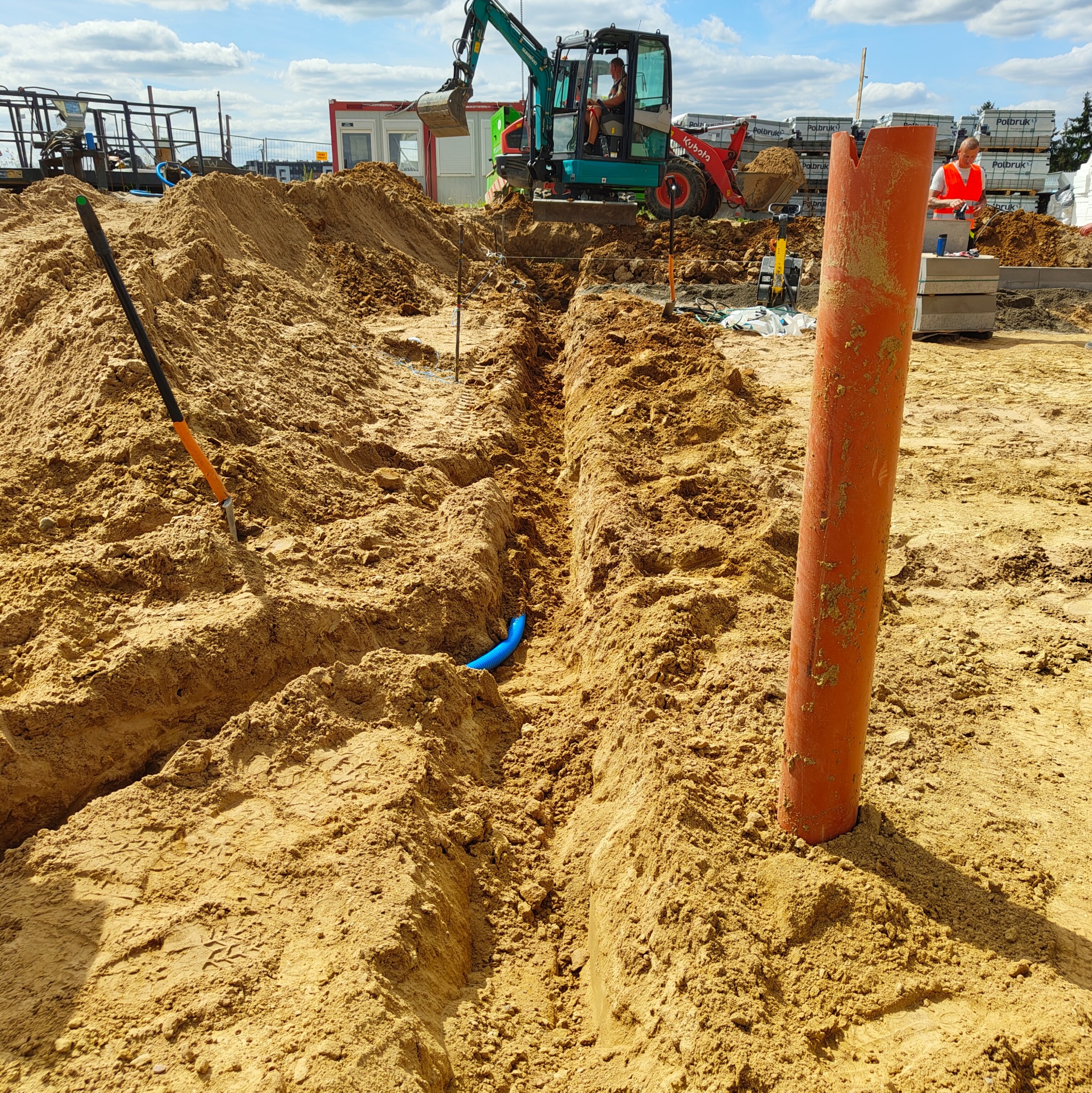 Wykopany rów w piasku z niebieską rurą w środku i pomarańczową rurą obok. W tle koparka i pracownik w kamizelce. Prace ziemne pod instalacje.