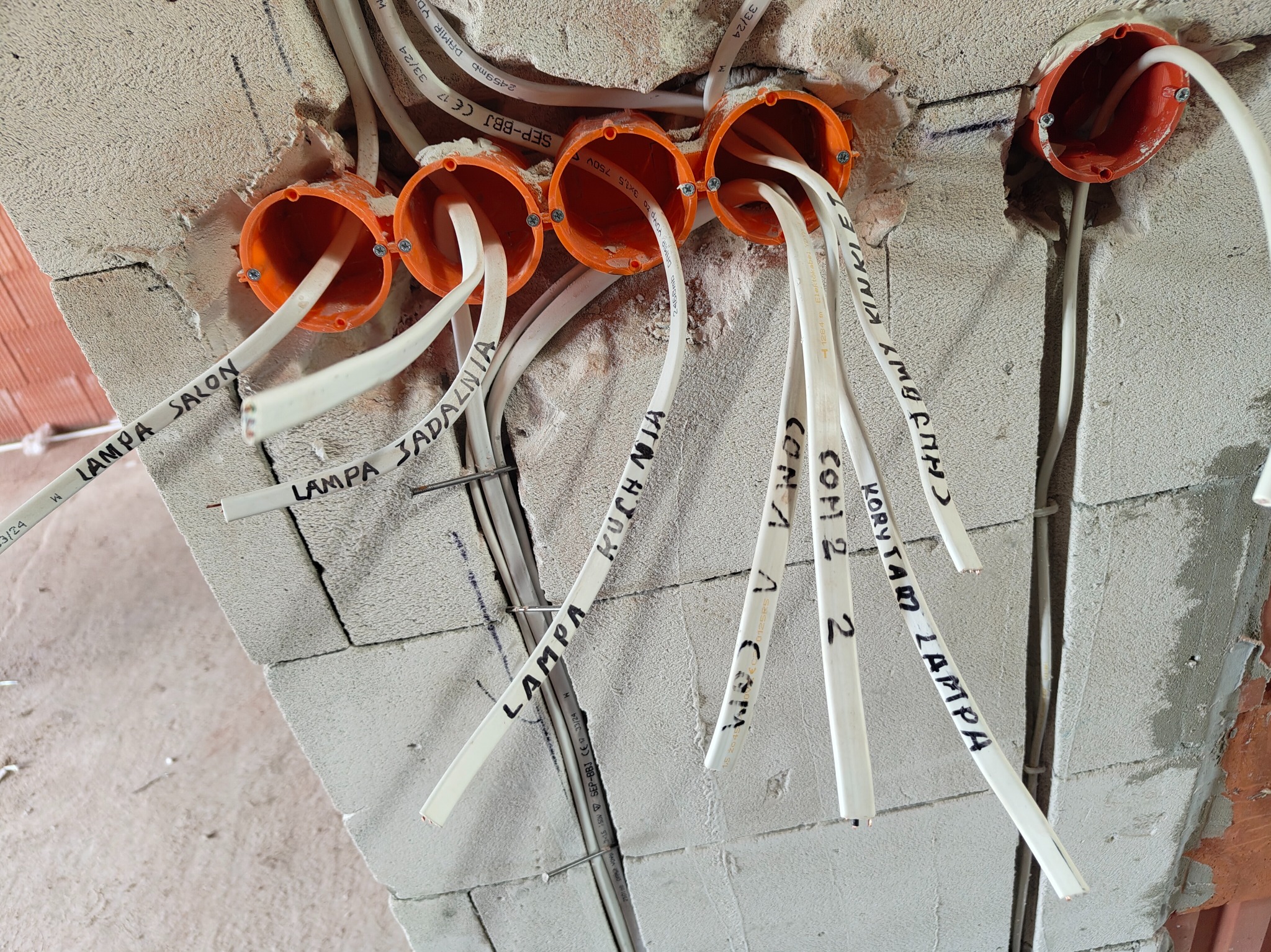 Pomarańczowe puszki elektryczne w surowej ścianie z wystającymi, opisanymi kablami: 'Lampa Salon', 'Lampa Jadalnia', 'Kuchnia', 'Korytarz Lampa'.