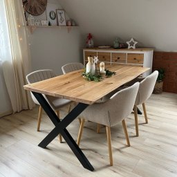 Sucha Decha - Stół z blatem z jasnego drewna i czarnymi nogami w kształcie litery X, otoczony czterema krzesłami w neutralnych odcieniach, w jasnym wnętrzu.