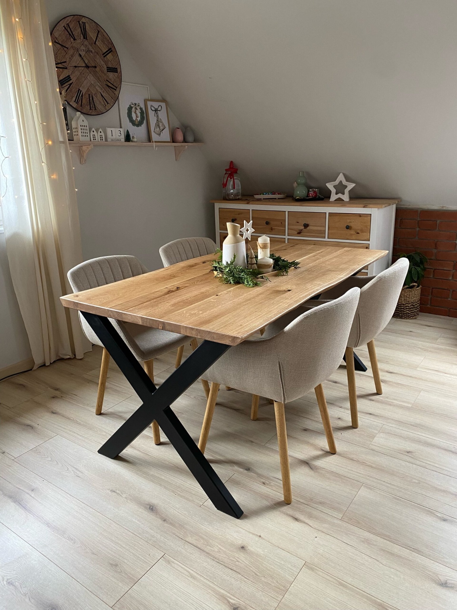 Stół z blatem z jasnego drewna i czarnymi nogami w kształcie litery X, otoczony czterema krzesłami w neutralnych odcieniach, w jasnym wnętrzu.