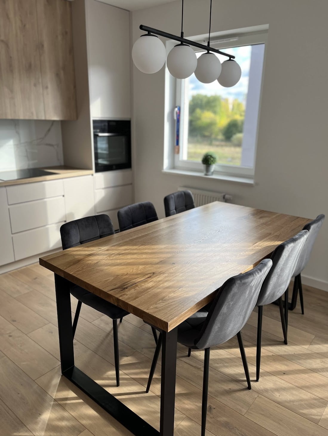 Stół z blatem drewnianym o wzorze jodełki, na czarnych, metalowych nogach, otoczony szarymi krzesłami, w nowoczesnej kuchni z oknem na zewnątrz.
