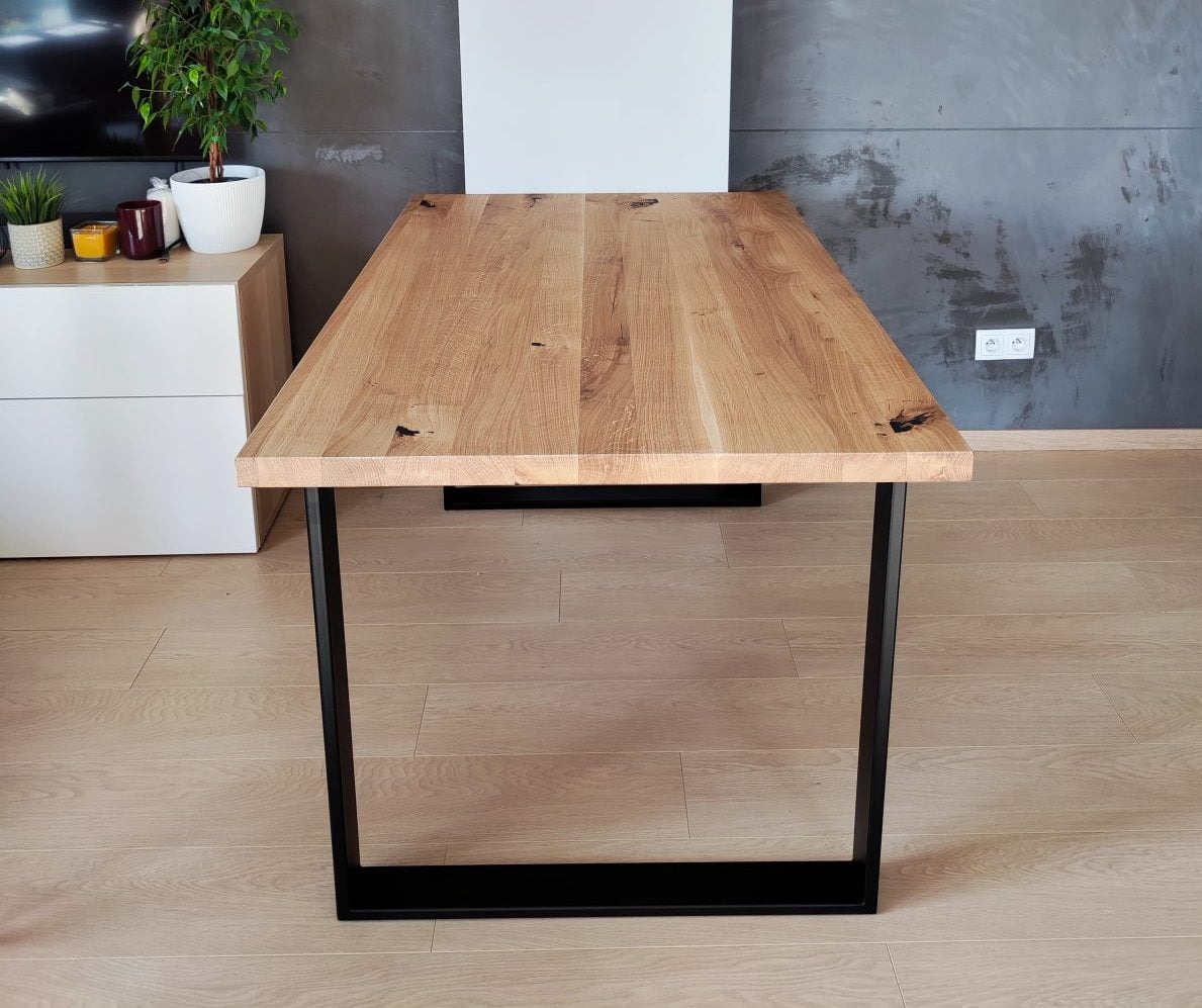 Stół z blatem z jasnego drewna o wyraźnym usłojeniu, na czarnych, metalowych nogach. W tle szara ściana i biała komoda z roślinami doniczkowymi.