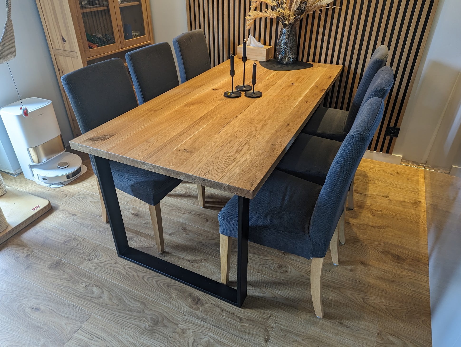 Stół z blatem z jasnego drewna i czarnymi nogami, otoczony szarymi krzesłami na tle drewnianej ściany. Na stole dekoracje: świeczniki, wazon z suszem. W tle widoczny robot sprzątający.