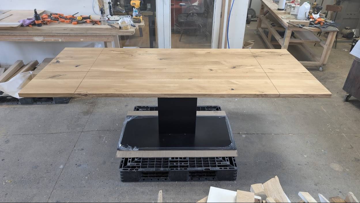 Blat drewniany z centralną nogą w trakcie montażu w Olsztynie. Widoczne łączenia desek i surowy charakter drewna. Całość na paletach, w tle warsztat.