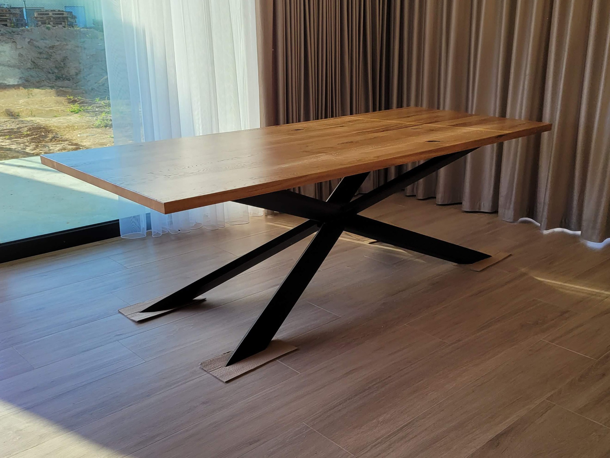 Stół z blatem z jasnego drewna i czarnymi, metalowymi nogami w kształcie litery X, ustawiony w jasnym wnętrzu z zasłonami i widokiem na zewnątrz.