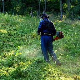BARTŁOMIEJ KASZAK USŁUGI LEŚNE - Mężczyzna w niebieskim kombinezonie kosi wysoką trawę na zboczu, używając profesjonalnej kosiarki spalinowej z osłoną. Widok z tyłu, w tle zieleń drzew.