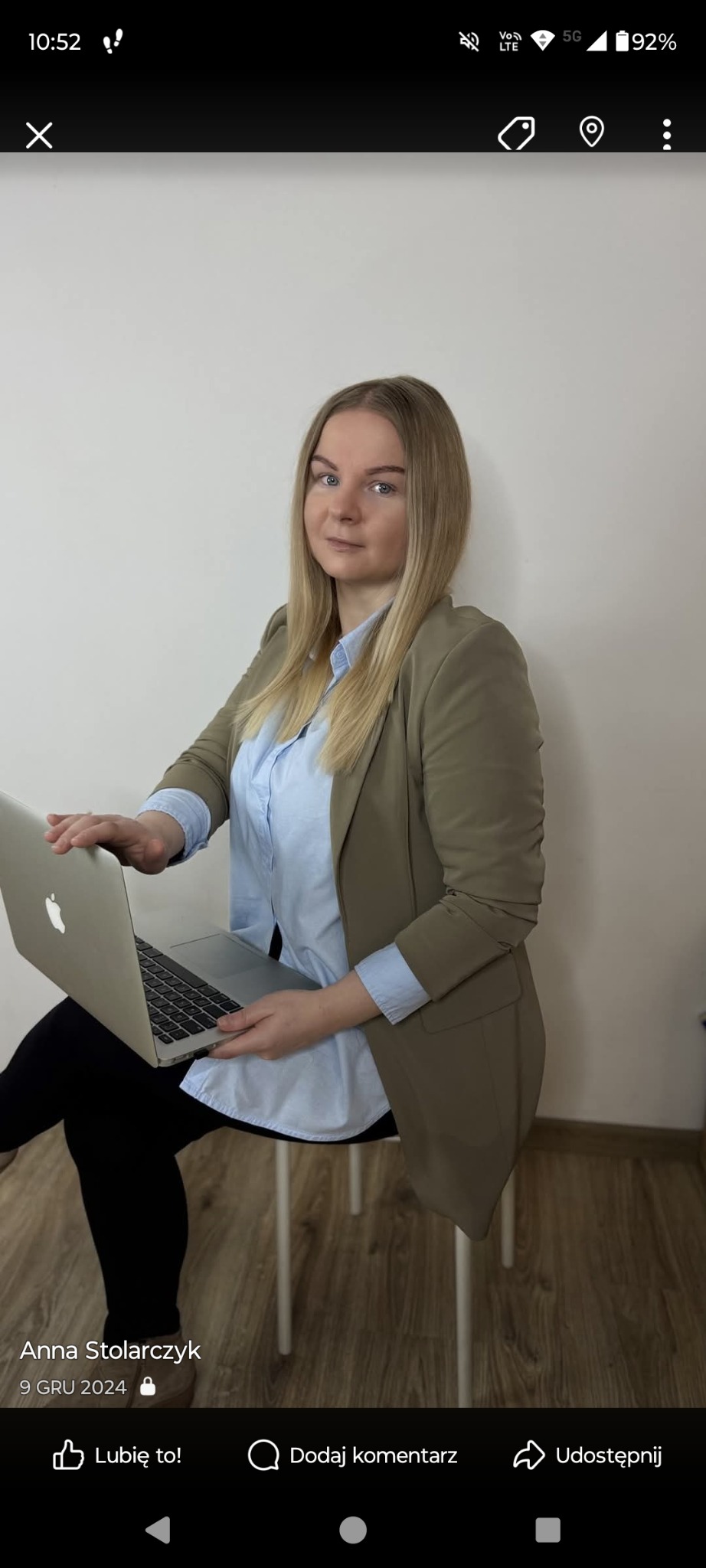 Kobieta z blond włosami w beżowej marynarce i błękitnej koszuli siedzi z laptopem na kolanach na białym krześle, pozując na tle jasnej ściany.