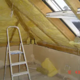 Ocieplenie poddasza wełną mineralną, widoczne drewniane krokwie, okno dachowe Fakro, aluminiowa drabina przystawna i fragment otynkowanej ściany.