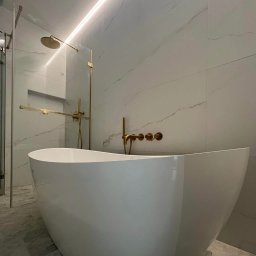 Łukasz Całka - Nowoczesna łazienka z wolnostojącą wanną, złotą armaturą i marmurowymi płytkami. Oświetlenie LED podkreśla elegancki design. W tle kabina prysznicowa.
