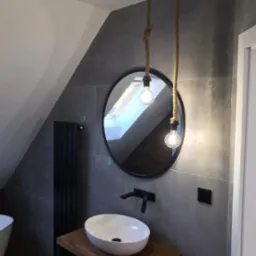 Nowoczesna łazienka na poddaszu z owalną umywalką na drewnianym blacie, okrągłym lustrem i lampami na sznurach. Szare płytki, czarne akcenty.