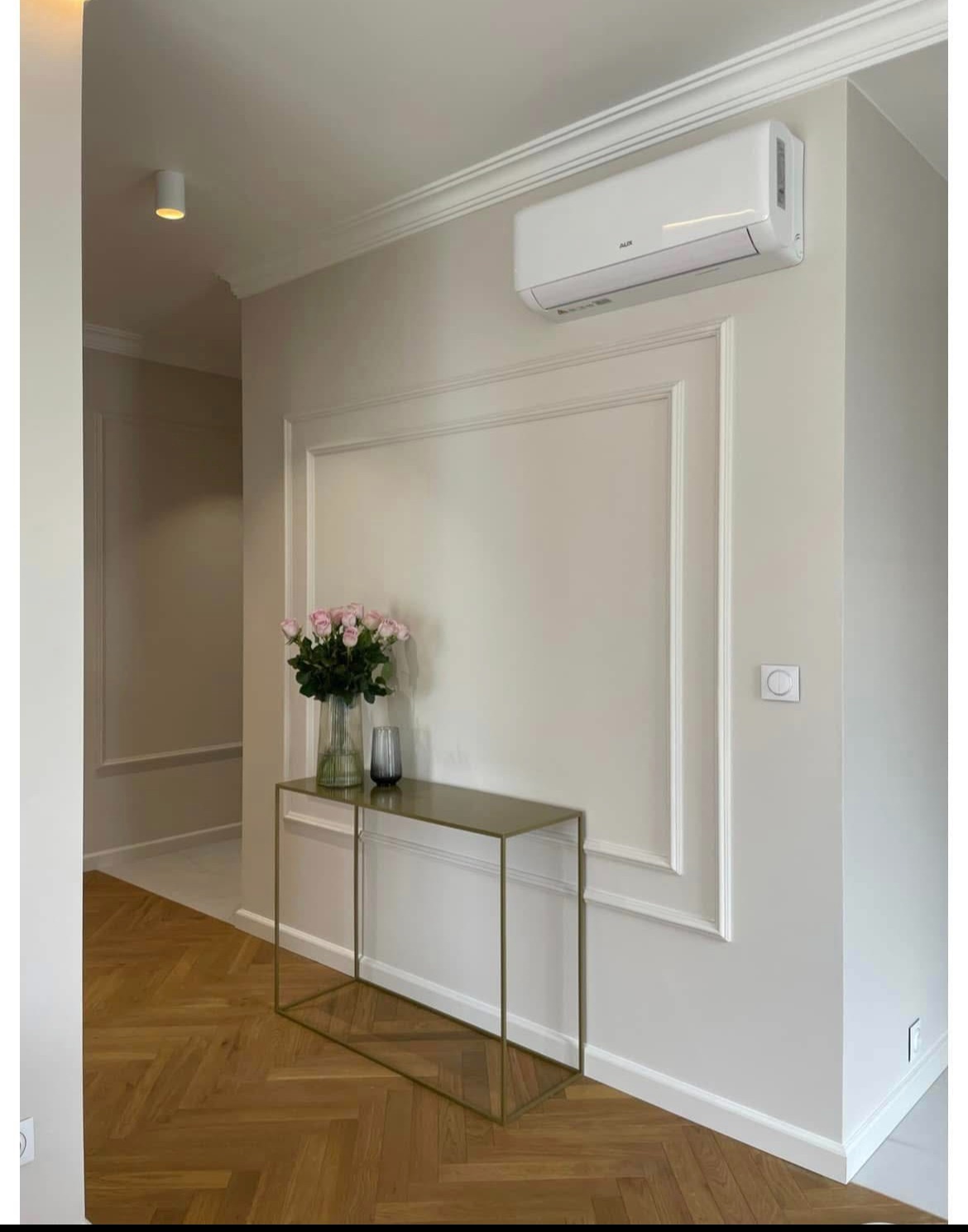 Elegancki przedpokój z parkietem w jodełkę, konsolą ze złotym stelażem, różami w wazonie i klimatyzatorem. Dekoracyjne listwy ścienne.