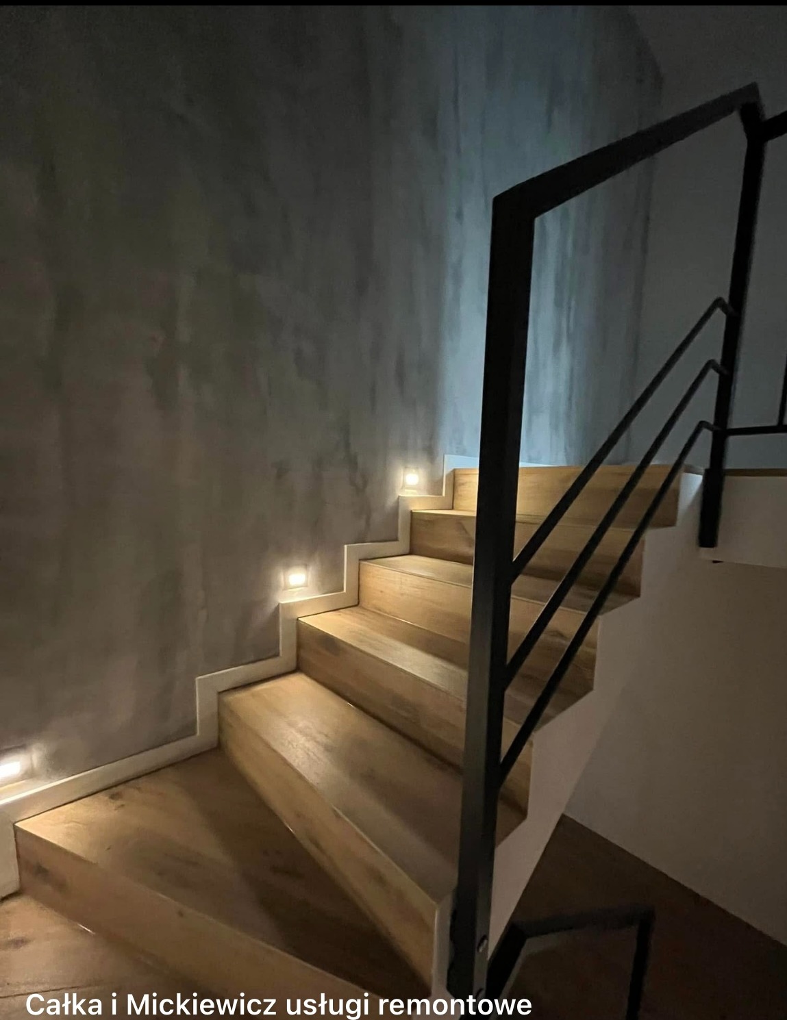 Nowoczesne schody drewniane z czarną balustradą i subtelnym oświetleniem LED w ścianie o fakturze betonu. Minimalistyczny design.