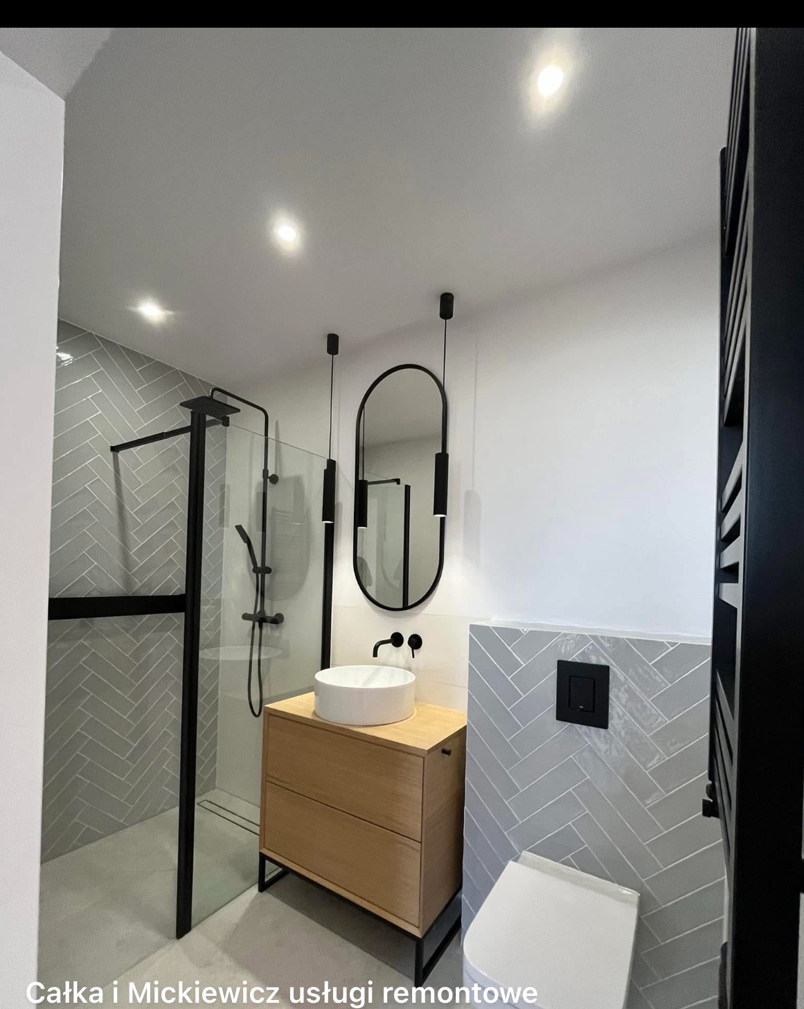 Nowoczesna łazienka po remoncie: kabina prysznicowa z czarnymi ramami, umywalka nablatowa na drewnianej szafce, lustro z oświetleniem i szare płytki ułożone w jodełkę.