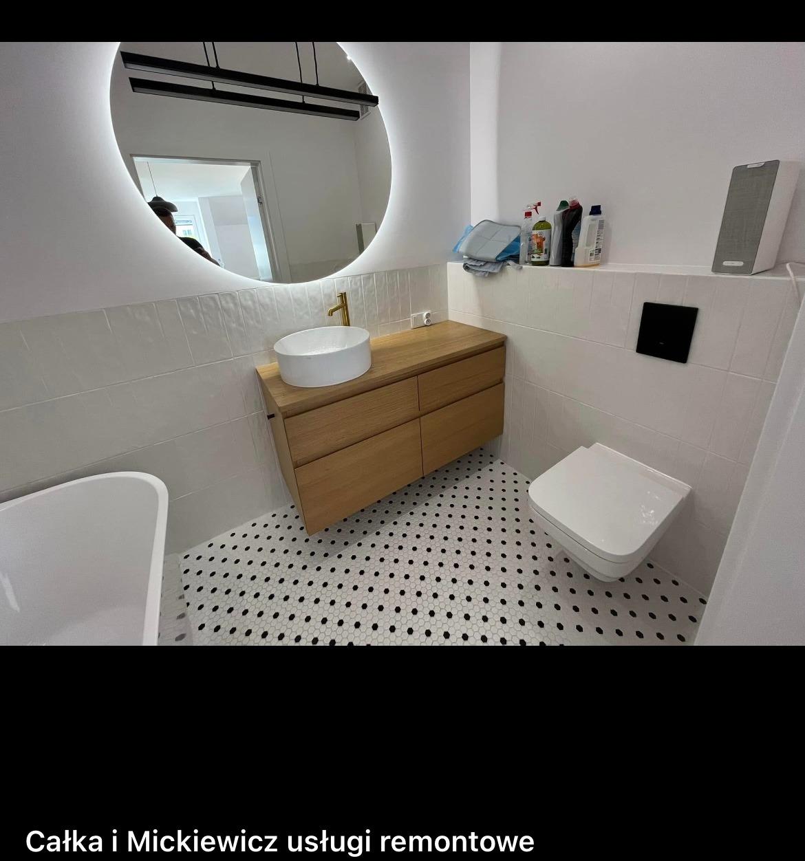 Nowoczesna łazienka po remoncie: białe płytki, drewniana szafka z umywalką, okrągłe lustro z podświetleniem LED i mozaika na podłodze w czarno-białe kropki.