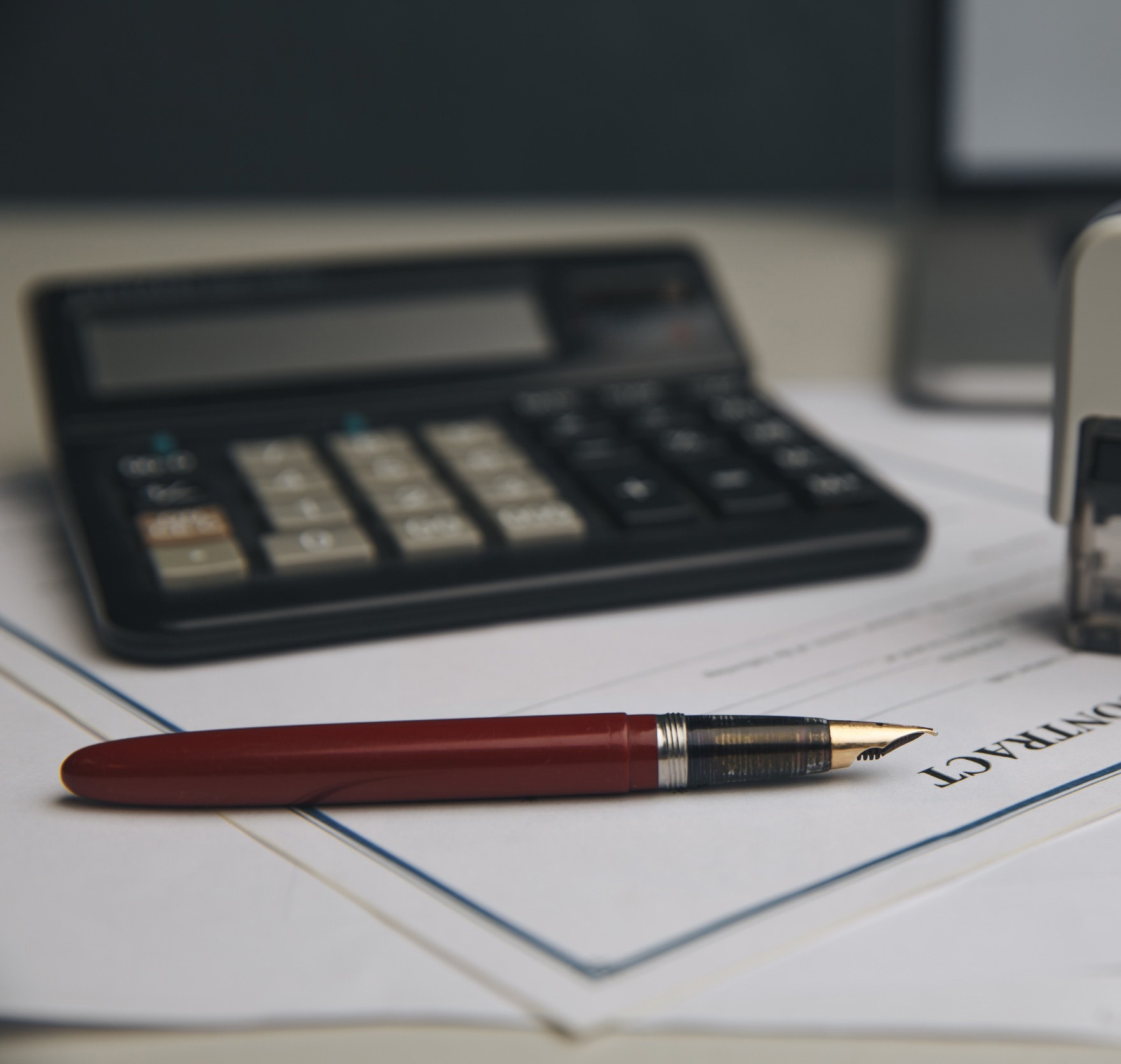 Umowa z piórem, kalkulatorem i pieczątką w tle. Ciemnoczerwone pióro wieczne leży na dokumencie. Elementy biurowe na jasnym blacie biurka.