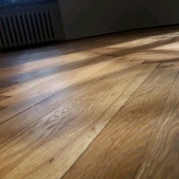 Prywatno - Parkiet ułożony w jodełkę, oświetlony naturalnym światłem z okna, z widocznym grzejnikiem żeliwnym w tle. Jasne drewno, ciepłe odcienie.