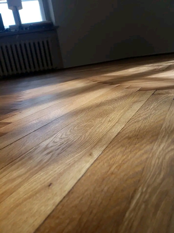 Parkiet w jodełkę, oświetlony promieniami słońca wpadającymi przez okno. Widoczny fragment grzejnika i ściany w tle. Podłoga po renowacji lub nowa.