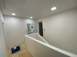 Białe, świeżo pomalowane wnętrze z drewnianą podłogą i widoczną balustradą. W tle, przez drzwi, widać fragment łazienki.  Oświetlenie sufitowe.