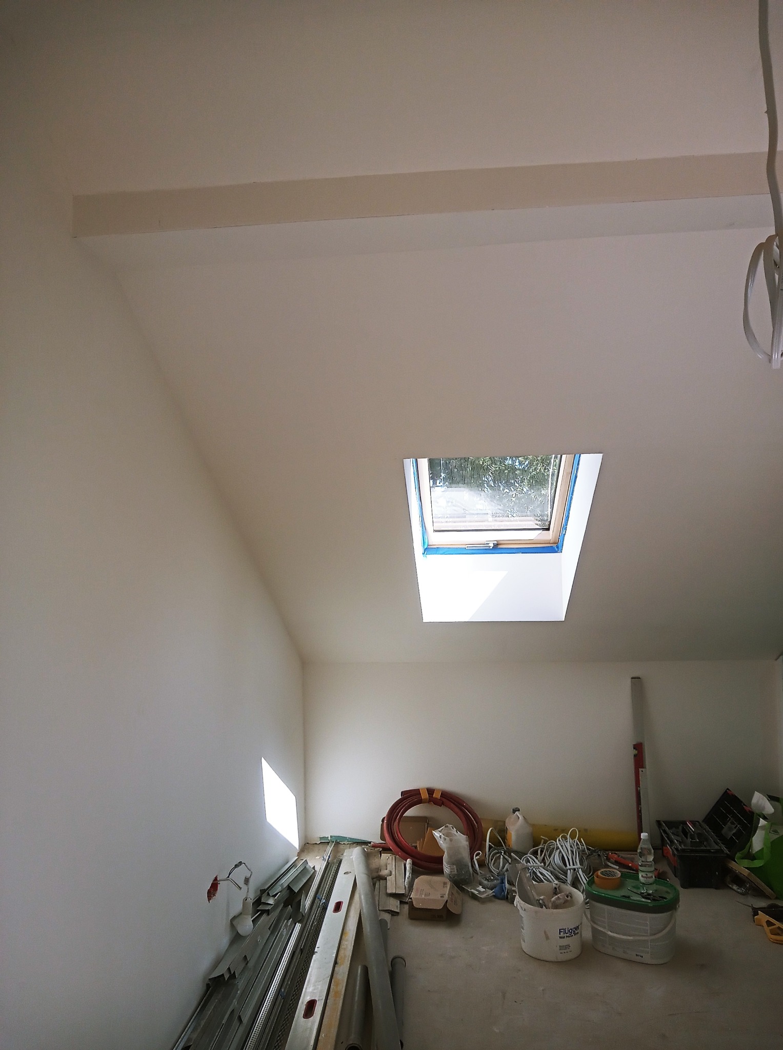 Poddasze po szpachlowaniu i malowaniu na biało, z oknem dachowym zabezpieczonym taśmą malarską. Na podłodze narzędzia i materiały budowlane. Widok z dołu.