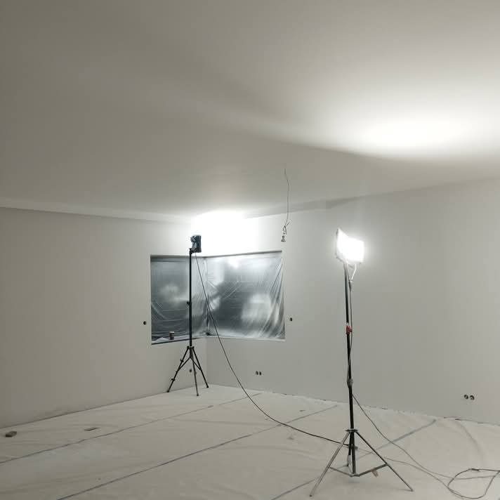 Białe, odświeżone wnętrze pokoju z osłoniętym oknem. Przygotowanie do malowania, widoczne lampy oświetleniowe i folia ochronna na podłodze.