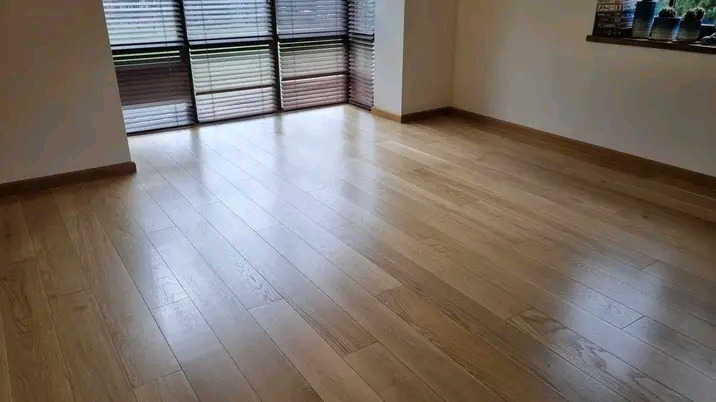 Lśniąca, parkietowa podłoga w nowocześnie wykończonym wnętrzu z dużym oknem z żaluzjami. Widoczne listwy przypodłogowe i parapet z dekoracjami.