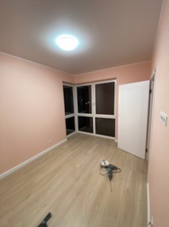 Pomalowane na różowo wnętrze pokoju po remoncie z jasną podłogą, widoczne narzędzia, otwarte drzwi i okno narożne z widokiem na nocne miasto.