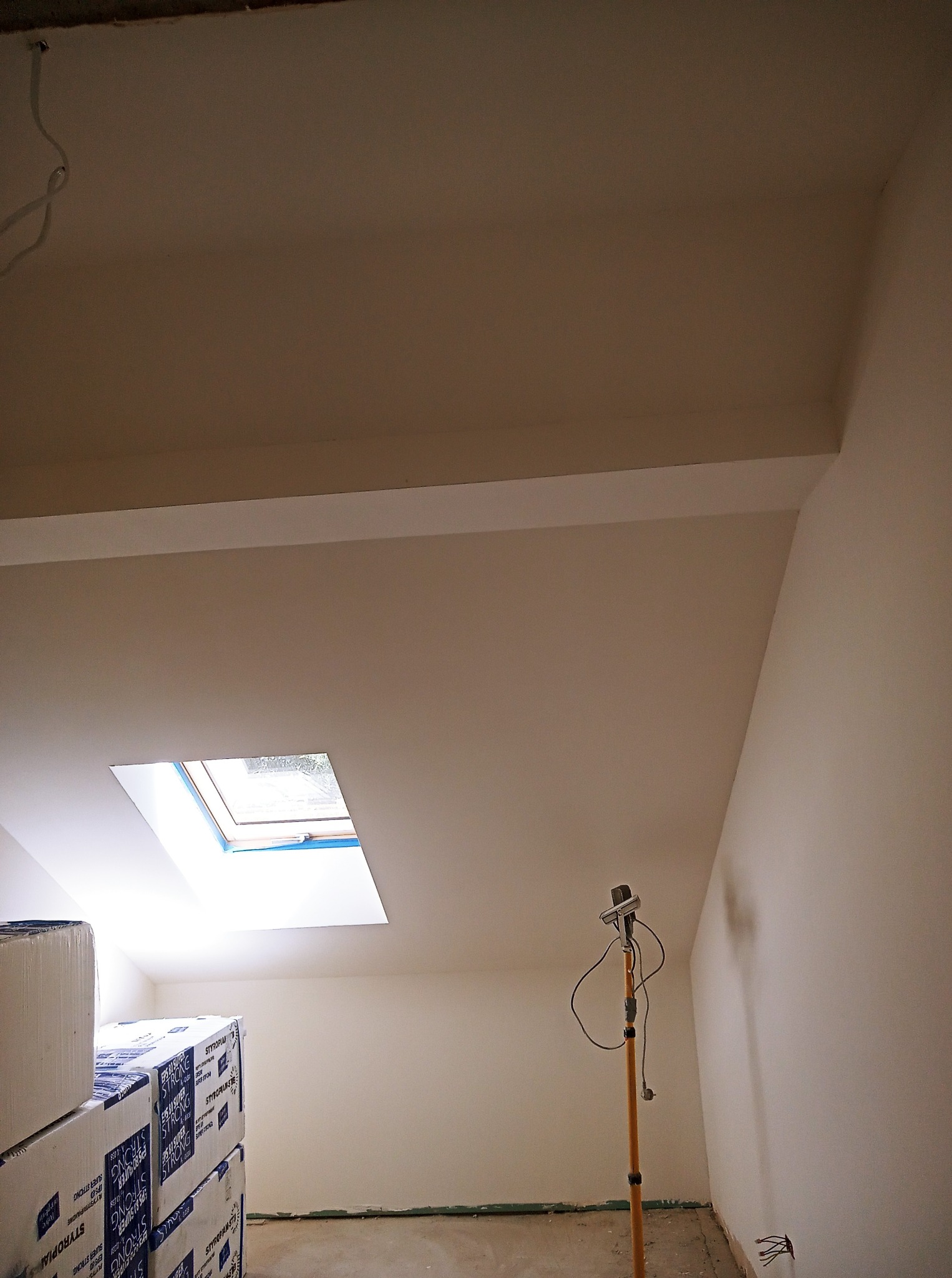 Poddasze po remoncie: świeżo pomalowane ściany w jasnym odcieniu, okno dachowe i materiały budowlane. Widoczny reflektor na statywie doświetlający wnętrze.