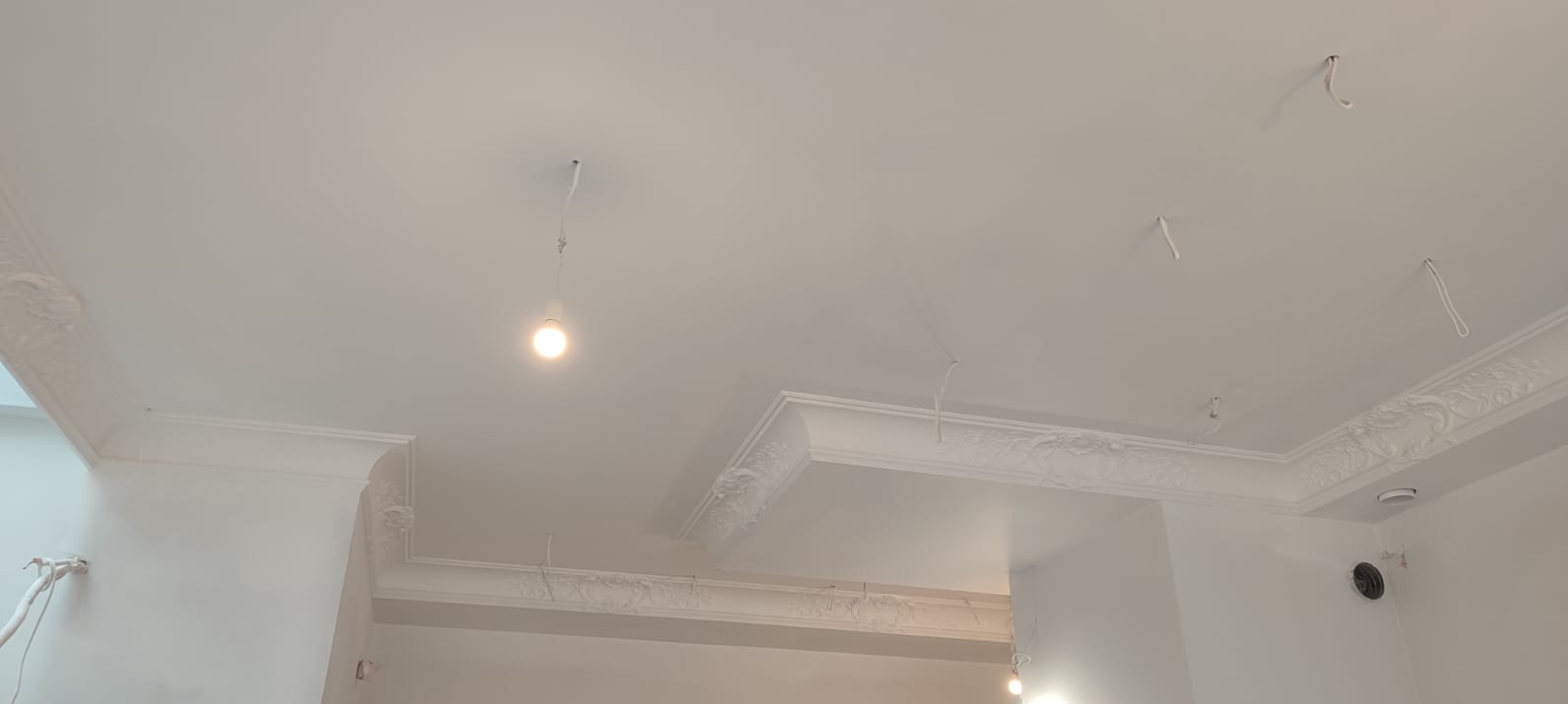 Biały sufit z dekoracyjnymi listwami gipsowymi, widoczne kable oświetleniowe, świeżo po malowaniu. Detal wykończenia wnętrza.