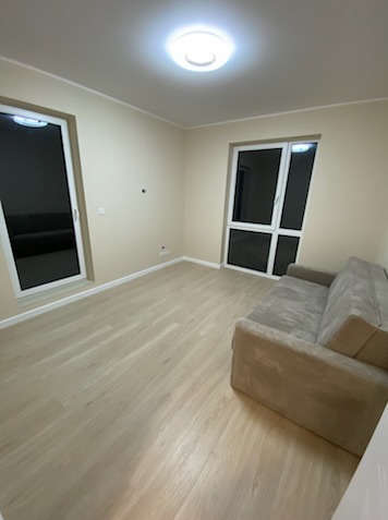 Jasny pokój po remoncie: kremowe ściany, drewniana podłoga, sofa i duże okna z białymi ramami. Sufit z okrągłą lampą. Minimalistyczny styl, neutralne kolory.