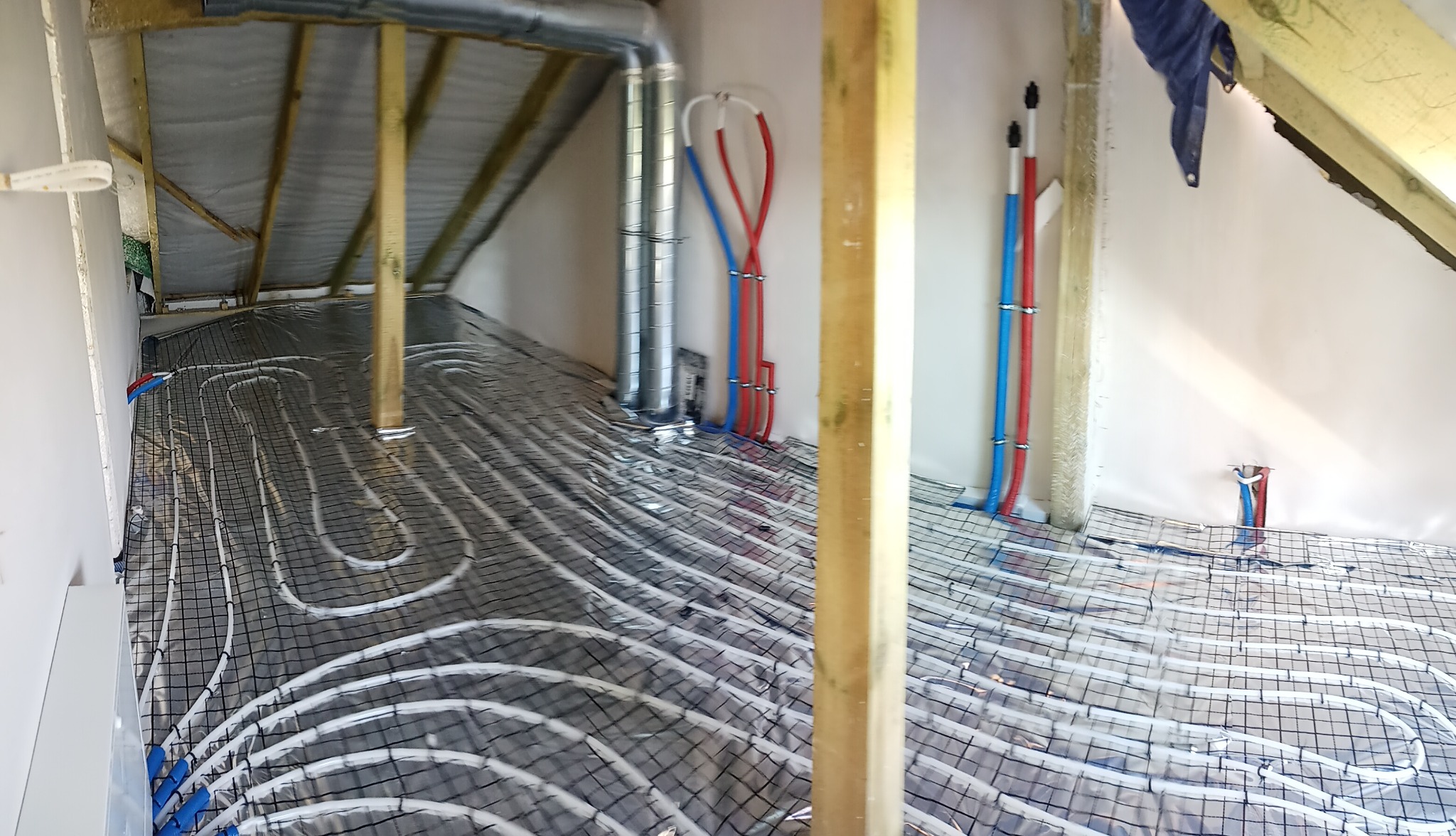 Instalacja ogrzewania podłogowego w poddaszu, rury ułożone na folii refleksyjnej, widoczne pionowe rury doprowadzające, drewniane elementy konstrukcyjne dachu.