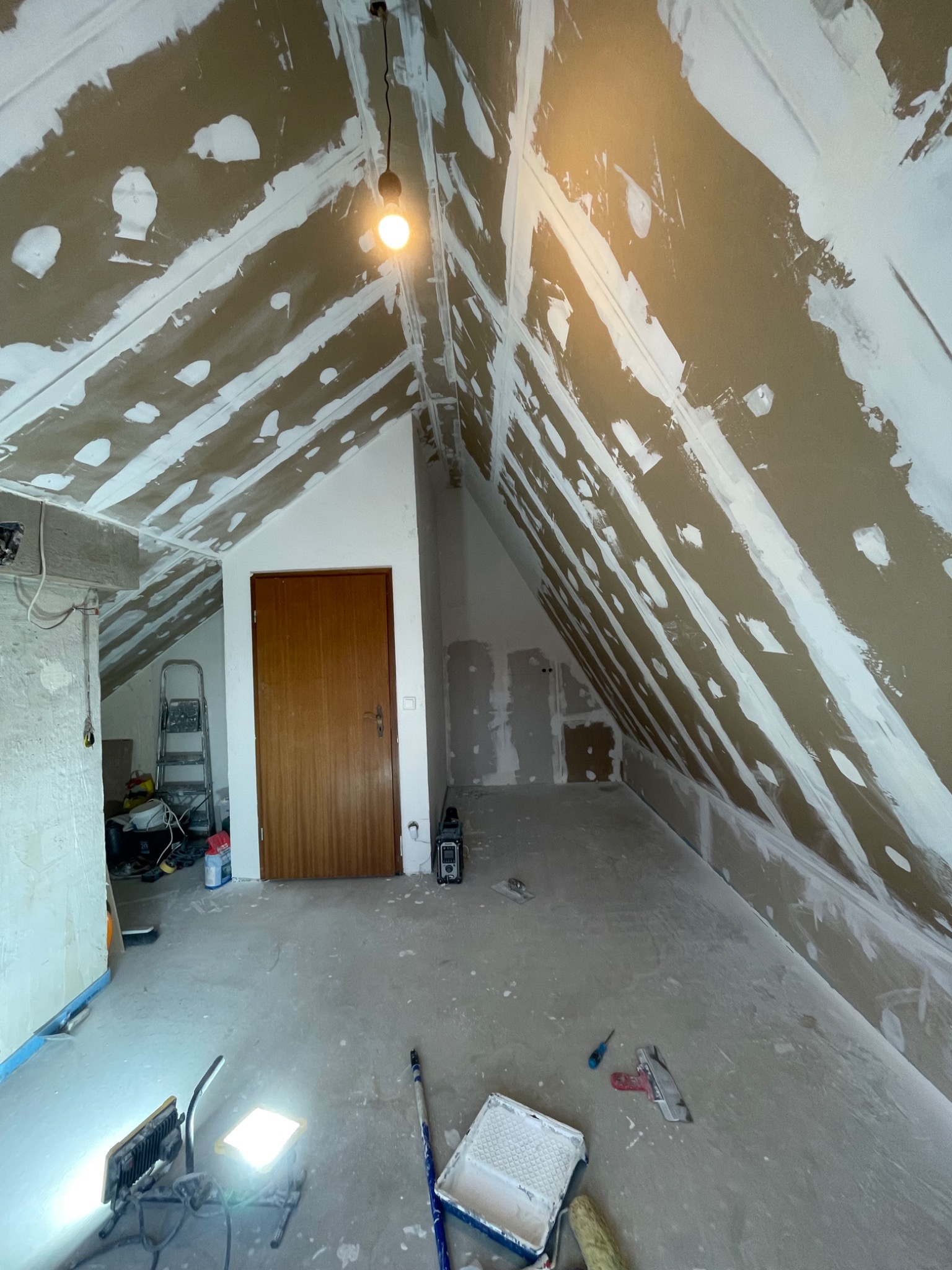 Poddasze w trakcie szpachlowania: widoczne pasy gładzi na skośnych ścianach i suficie, narzędzia malarskie na podłodze, surowa żarówka. Etap prac wykończeniowych.