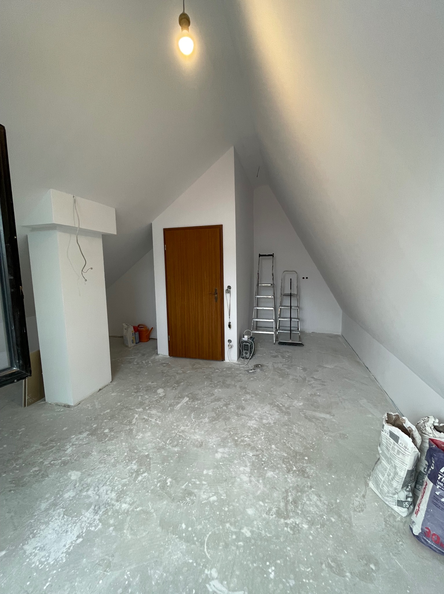 Białe poddasze po szpachlowaniu, widoczne worki z zaprawą, drabiny i świeżo pomalowane ściany. Przygotowanie do dalszych prac wykończeniowych.