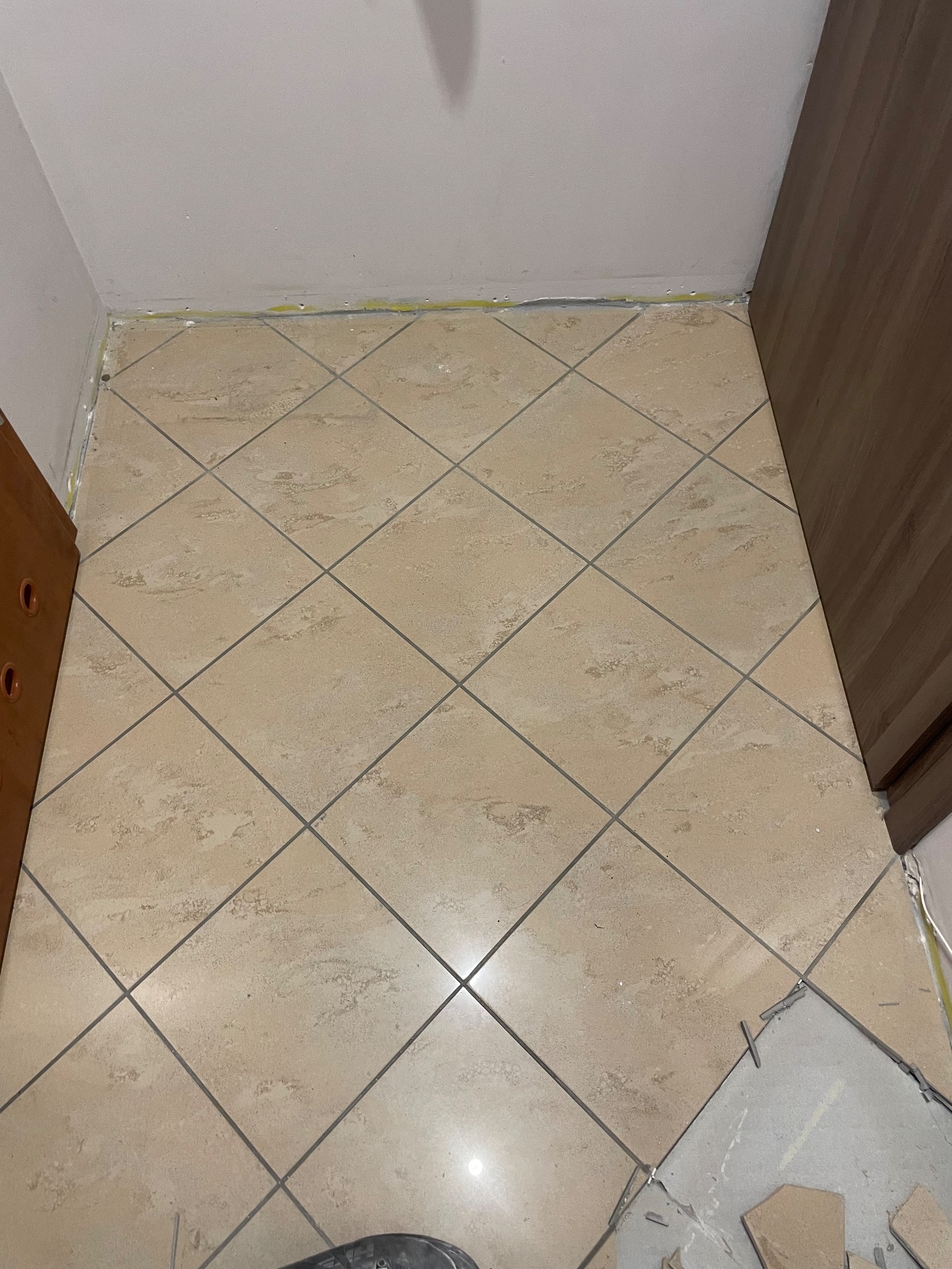 Beżowe płytki podłogowe ułożone w karo, widoczne fragmenty niedokończonej pracy przy krawędzi ściany i szafki. Żółta taśma malarska przy suficie.