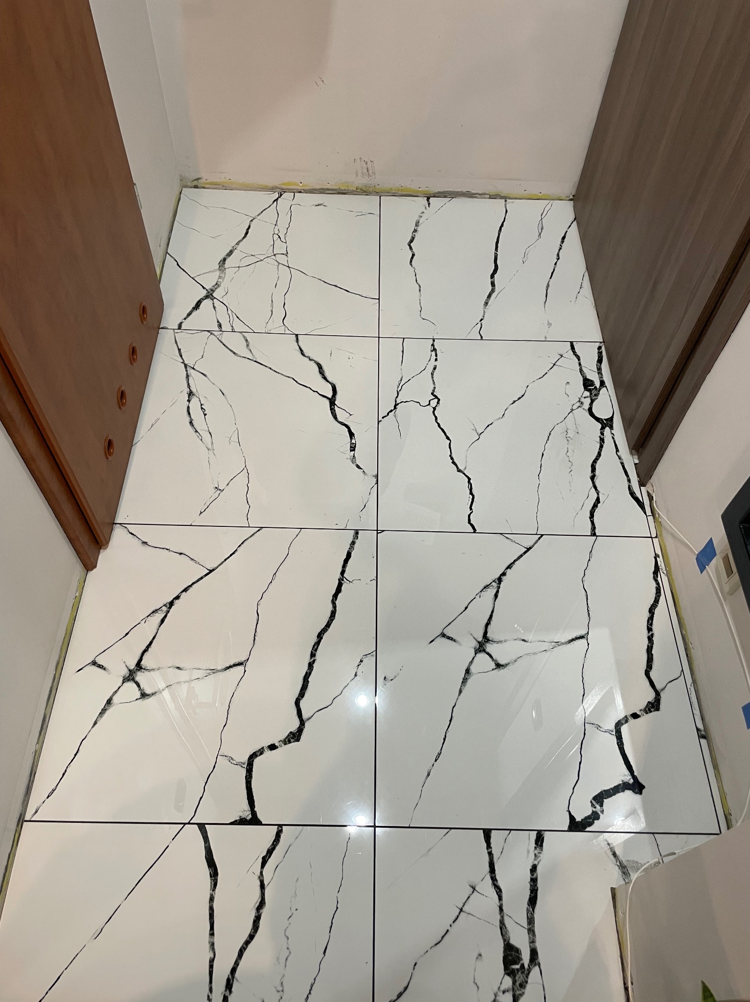 Błyszczące, białe płytki podłogowe z czarnym marmurkowym wzorem, ułożone w wąskim korytarzu między brązowymi szafkami. Widoczne fugi i detale wykończenia.