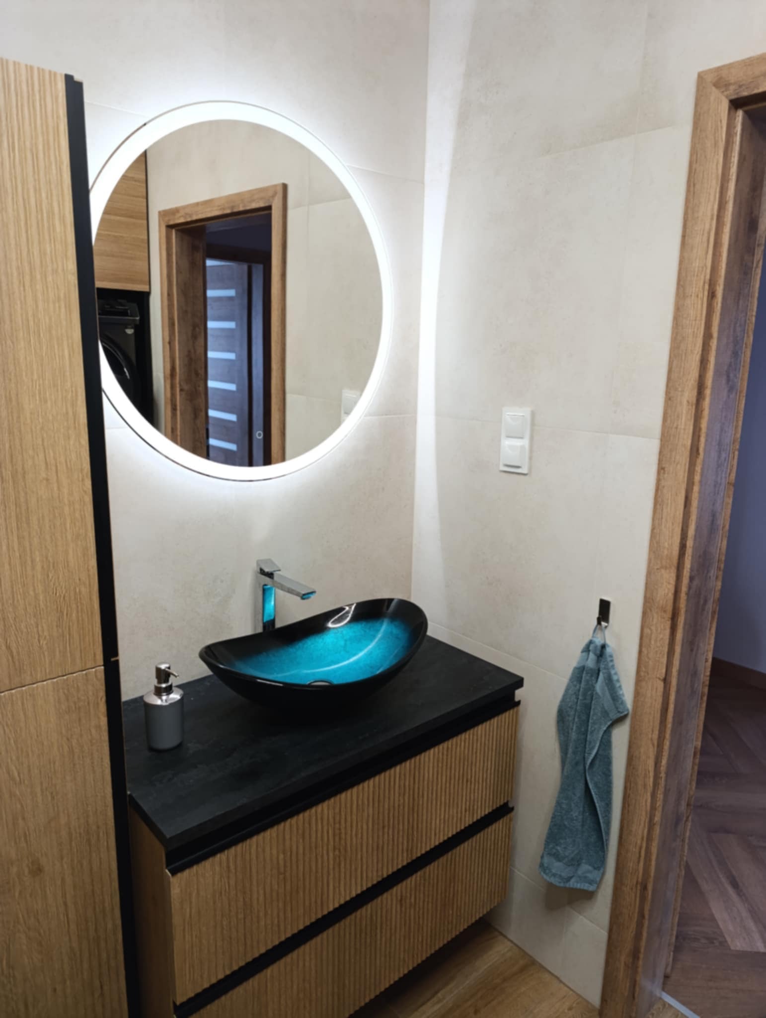 Nowoczesna łazienka: szafka z umywalką w kolorze turkusowym, okrągłe lustro z podświetleniem LED, beżowe płytki i drewniana podłoga.