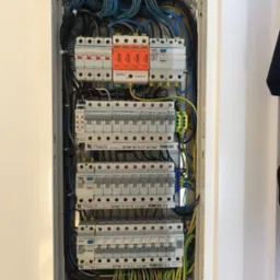 Rozdzielnica elektryczna z bezpiecznikami, przewodami i listwami zaciskowymi. Widoczne elementy marki Hager i Simtec. Całość w białej obudowie.